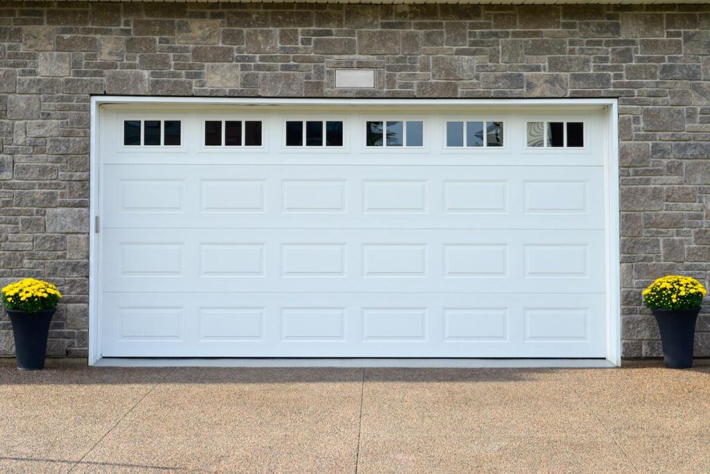 A large white metal panel residential garage door with small glass windows on a grey brick building.There are two pots with yellow flowers on either side of the door. The ground is a beige block drive