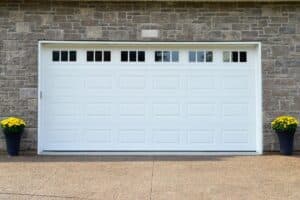 A large white metal panel residential garage door with small glass windows on a grey brick building.There are two pots with yellow flowers on either side of the door. The ground is a beige block drive